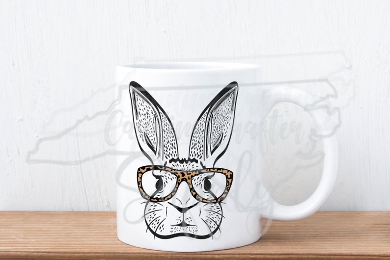 May include: White ceramic mug featuring a black and white illustration of a rabbit wearing leopard print glasses. The rabbit has long ears and a detailed face. The mug has a white handle and is set against a light background.