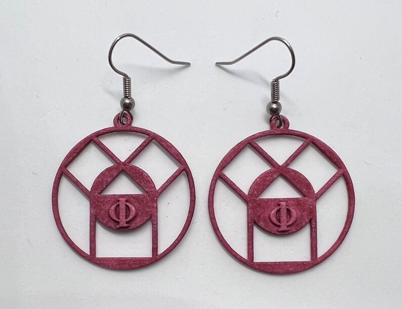 May include: A pair of pink earrings with a geometric design featuring a circle with a triangle inside and the Greek letter Phi in the center. The earrings have silver hooks.
