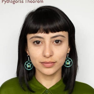 May include: A woman with black hair and bangs wearing a green shirt and turquoise earrings. The earrings are round and have a geometric design.