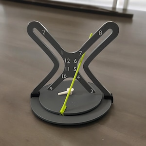 May include: A grey clock with a unique design featuring a circular face with numbers 1 through 12 and a yellow hand. The clock is mounted on a grey X-shaped stand.