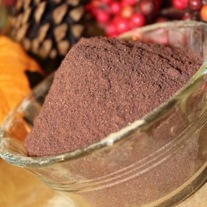 May include: A glass bowl filled with a mound of dark brown powder. The powder is a deep reddish brown color and has a fine texture. The bowl is sitting on a wooden surface.