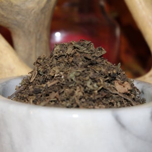 May include: A white marble mortar and pestle filled with dried herbs. The herbs are a dark brown color and have a rough texture.