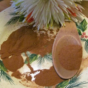 Op de afbeelding: Een witte en rode bloem met een houten lepel gevuld met bruin kaneelpoeder op een wit bord met een groen en bruin bloemenontwerp.
