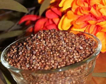 May include: A glass bowl filled with brown and white peppercorns. The bowl is sitting on a wooden surface. There are orange and red flowers in the background.