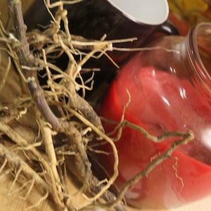 May include: A close-up of a red candle in a glass jar, surrounded by dried roots and branches on a wooden surface.