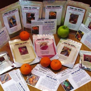 May include: A variety of organic spice and tea blends in clear plastic bags with labels that say "Tame the Spirit" and list the ingredients. The bags are arranged on a wooden table with green apples and oranges.