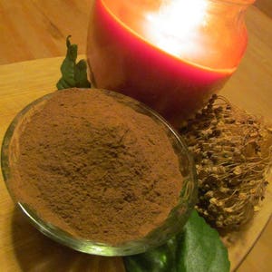 May include: A close-up of a small glass bowl filled with a brown powder, a green leaf, and a dried flower arrangement. A red candle in a glass jar is out of focus in the background.