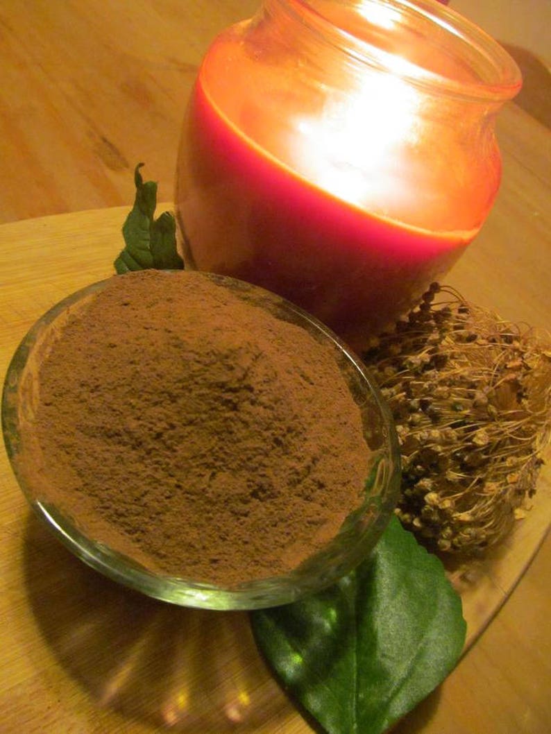 May include: A close-up of a small glass bowl filled with a brown powder, a green leaf, and a dried flower arrangement. A red candle in a glass jar is out of focus in the background.
