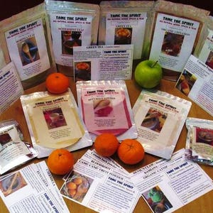 Puede incluir: Una variedad de especias y t&eacute;s envasados en peque&ntilde;as bolsas con etiquetas que dicen "Tame the Spirit". Las bolsas est&aacute;n dispuestas sobre una mesa de madera con naranjas y manzanas alrededor.