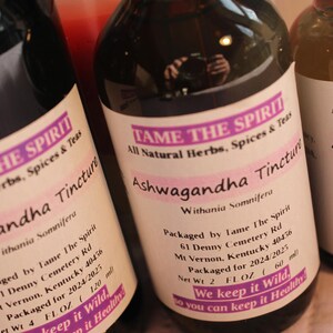 May include: Three glass bottles with brown liquid and white labels. The labels have the text "TAME THE SPIRIT" and "Ashwagandha Tincture" with details about the product and packaging. The bottles are on a white surface.