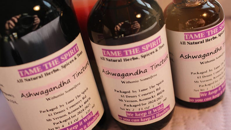 May include: Three glass bottles with brown liquid and white labels. The labels have the text "TAME THE SPIRIT" and "Ashwagandha Tincture" with details about the product and packaging. The bottles are on a white surface.