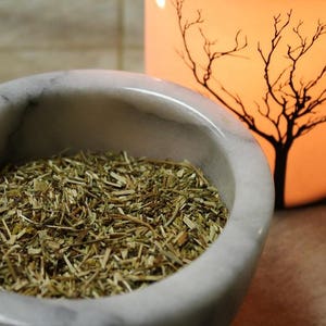 May include: A white marble mortar and pestle filled with dried herbs. The mortar is round and has a smooth surface. The herbs are a light brown color and are finely chopped.