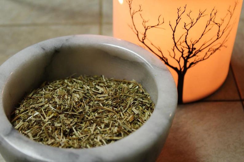 May include: A white marble mortar and pestle filled with dried herbs. The mortar is round and has a smooth surface. The herbs are a light brown color and are finely chopped.