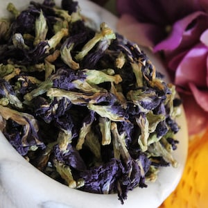 May include: A white bowl filled with dried blue butterfly pea flowers. The flowers are a vibrant blue with green stems.