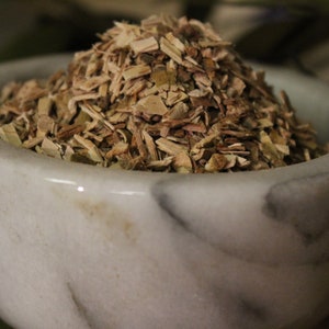 May include: A white marble mortar filled with a pile of dried, chopped herbs. The herbs are a mix of light brown and green, with a textured appearance. The mortar has a smooth, polished surface.