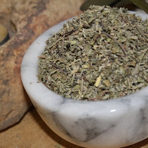 May include: A white marble bowl filled with dried green herbs. The bowl is sitting on a wooden surface.