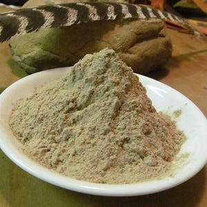 May include: A white ceramic dish filled with a mound of light brown powder. The powder is piled high in the center of the dish, creating a peak.