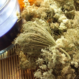 May include: A close-up of a purple candle in a clear glass container with three wicks. Dried, beige-colored flowers are arranged next to the candle on a bamboo mat. The candle and flowers create a relaxing, spa-like atmosphere.