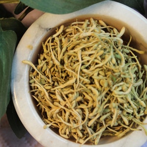 May include: A white ceramic bowl filled with dried, light brown flower buds. The bowl is sitting on a white surface.