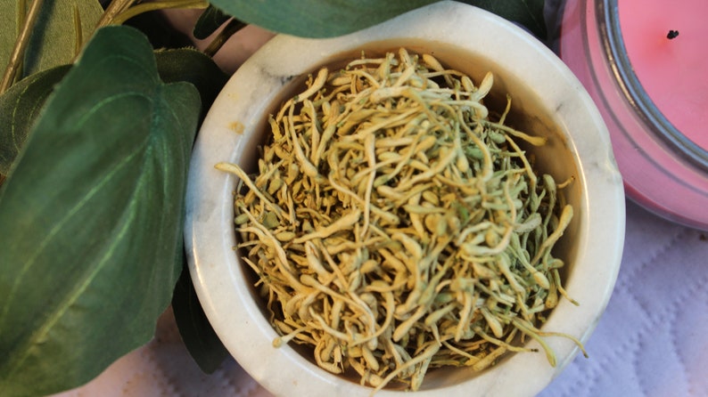 May include: A white ceramic bowl filled with dried, light brown flower buds. The bowl is sitting on a white surface.