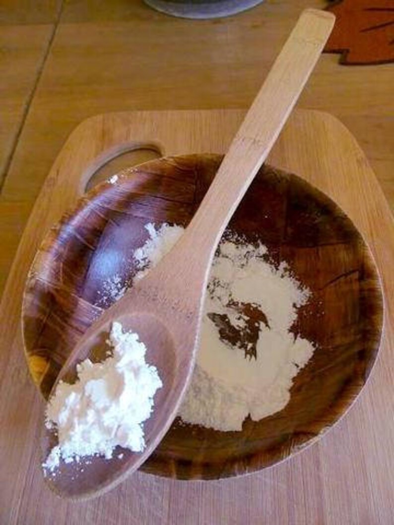 May include: A wooden spoon filled with white powder sits in a wooden bowl with more powder. The bowl and spoon are on a wooden surface.