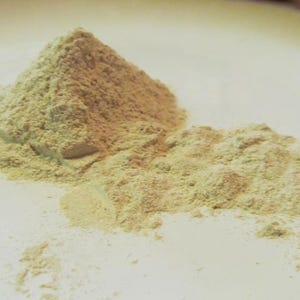Puede incluir: Un mont&oacute;n de polvo beige claro sobre una superficie blanca. El polvo est&aacute; finamente molido y tiene una textura ligeramente granulada.