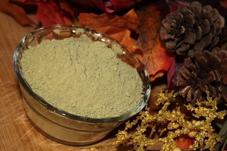 May include: A close-up of a small glass bowl filled with a light green powder. The bowl is sitting on a wooden surface with fall-colored leaves and pine cones around it.