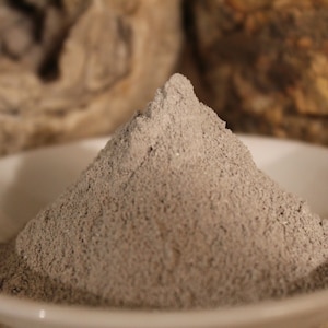 May include: A small pile of gray powder in a white ceramic bowl. The powder is finely ground and has a smooth texture.