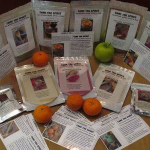 Op de afbeelding: Een assortiment kruidenproducten van "Tame the Spirit" wordt getoond met groene appels en sinaasappels. De producten zitten in doorzichtige zakjes met bedrukte labels met productnamen en afbeeldingen. De tekst "Tame the Spirit" is zichtbaar.