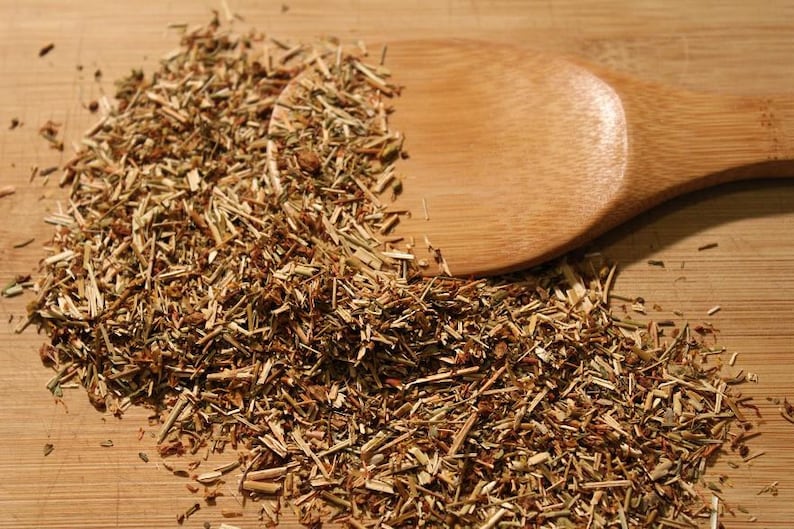 May include: A close-up of a pile of dried herbs on a wooden surface. A wooden spoon is lying on top of the herbs.