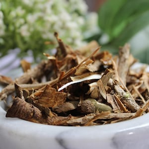 May include: Close-up of a white marble bowl filled with dried herbs and spices. The mixture includes brown, tan, and green pieces. White flowers and green leaves are visible in the background.