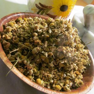 May include: A wooden bowl filled with dried chamomile flowers. The flowers are a light brown color and have a slightly yellow tint. The bowl is made of dark wood and has a natural finish.