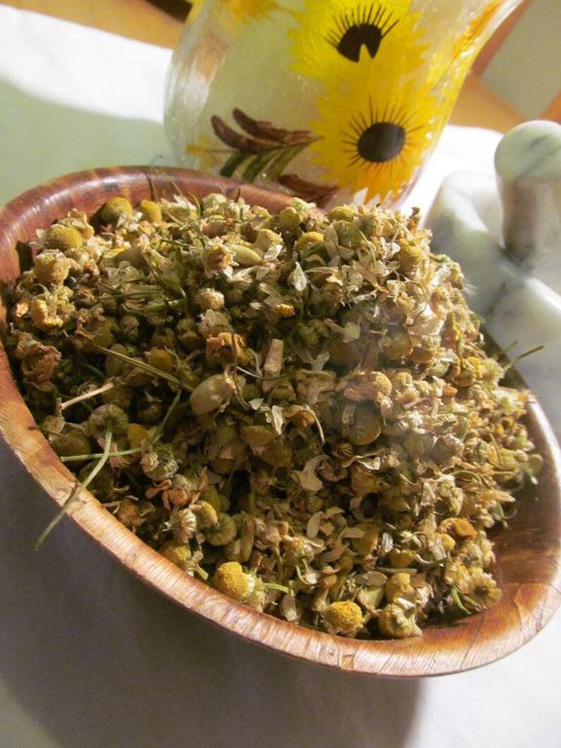 May include: A wooden bowl filled with dried chamomile flowers. The flowers are a light brown color and have a slightly yellow tint. The bowl is made of dark wood and has a natural finish.