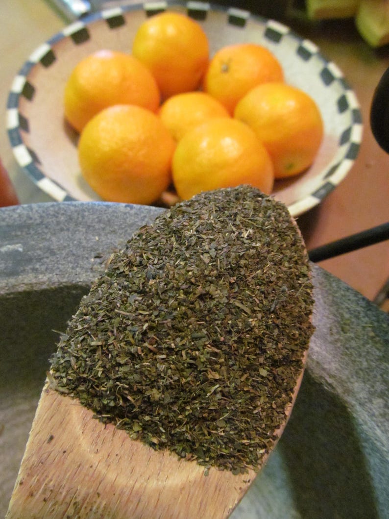 May include: A wooden spoon filled with dried green tea leaves. The spoon is sitting on top of a gray stone mortar. A bowl of oranges is in the background.
