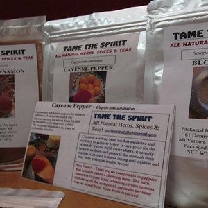 May include: Three bags of spices and herbs labeled "Tame The Spirit" with the names "Organic Cinnamon", "Cayenne Pepper", and "Bloodroot". The bags are on a wooden surface with apples in the foreground.