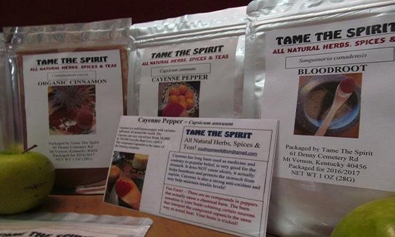 May include: Three bags of spices and herbs labeled "Tame The Spirit" with the names "Organic Cinnamon", "Cayenne Pepper", and "Bloodroot". The bags are on a wooden surface with apples in the foreground.