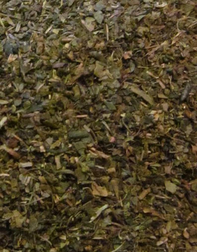 May include: Close-up of a pile of loose leaf green tea. The tea leaves are a mix of dark green and brown, with some small pieces of stem.