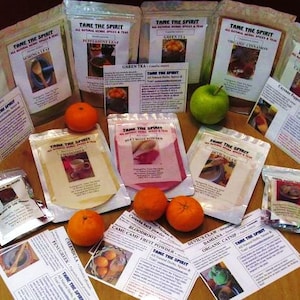 May include: A variety of spices and teas packaged in small bags with labels that say "Tame the Spirit". The bags are arranged on a wooden table with oranges and apples around them.