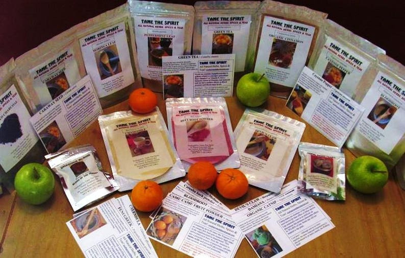 May include: A variety of spices and teas packaged in small bags with labels that say "Tame the Spirit". The bags are arranged on a wooden table with oranges and apples around them.