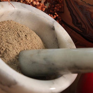 May include: A white marble mortar and pestle with a light brown powder inside. The mortar is partially filled with the powder.