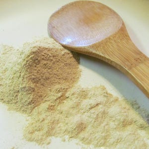 Puede incluir: Una cuchara de madera se encuentra sobre una superficie blanca junto a un mont&oacute;n de polvo marr&oacute;n claro. El polvo es probablemente una especia o una hierba.