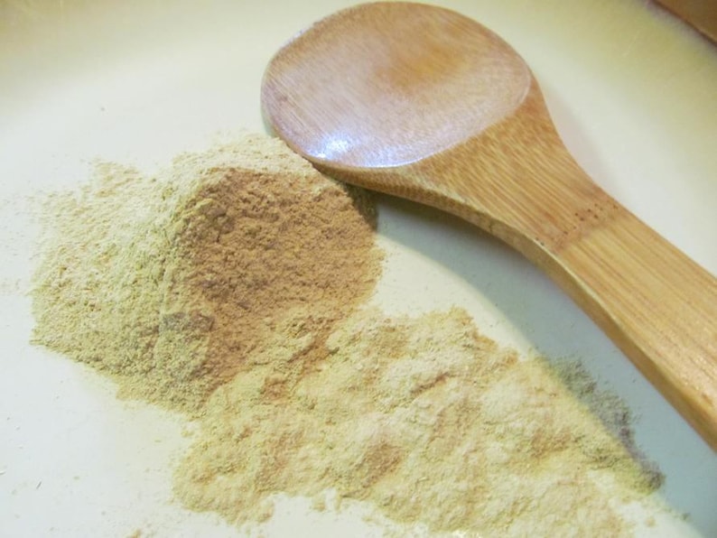 Puede incluir: Una cuchara de madera se encuentra sobre una superficie blanca junto a un mont&oacute;n de polvo marr&oacute;n claro. El polvo es probablemente una especia o una hierba.