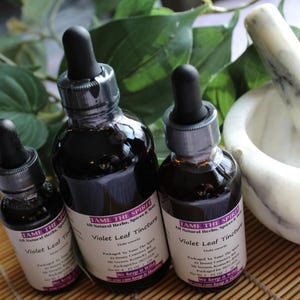 May include: Three dark glass bottles with droppers, filled with a dark liquid, labeled "Violet Leaf Tincture." A white marble mortar and pestle are on the right, with green leaves in the background. The bottles are on a bamboo mat.