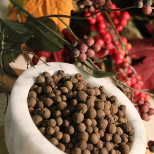 May include: A white marble bowl filled with whole allspice berries. The bowl is sitting on a wooden surface.