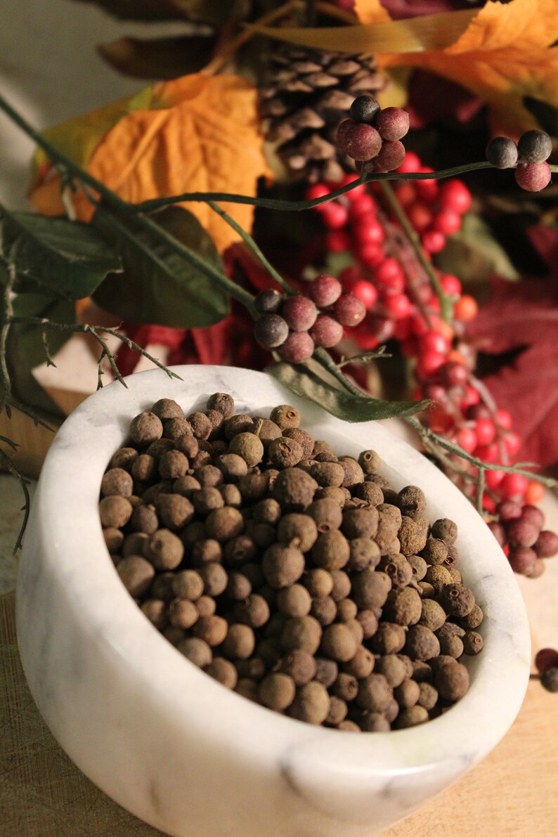 May include: A white marble bowl filled with whole allspice berries. The bowl is sitting on a wooden surface.