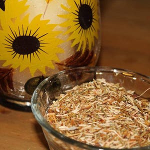 May include: A clear glass bowl filled with dried herbs, sitting on a wooden surface. The bowl is next to a glass vase with a sunflower design.
