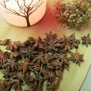 May include: A close-up of a pile of star anise pods on a wooden surface. The pods are brown and have a star shape. There is a candle holder with a tree design in the background.