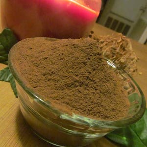 May include: A glass bowl filled with a heaping amount of dark brown powder. The powder is finely ground and has a smooth, even texture. The bowl is sitting on a wooden surface.