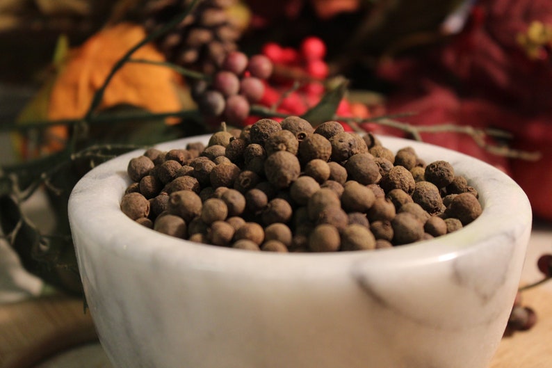 May include: A white ceramic bowl filled with whole allspice berries. The bowl is sitting on a wooden surface.