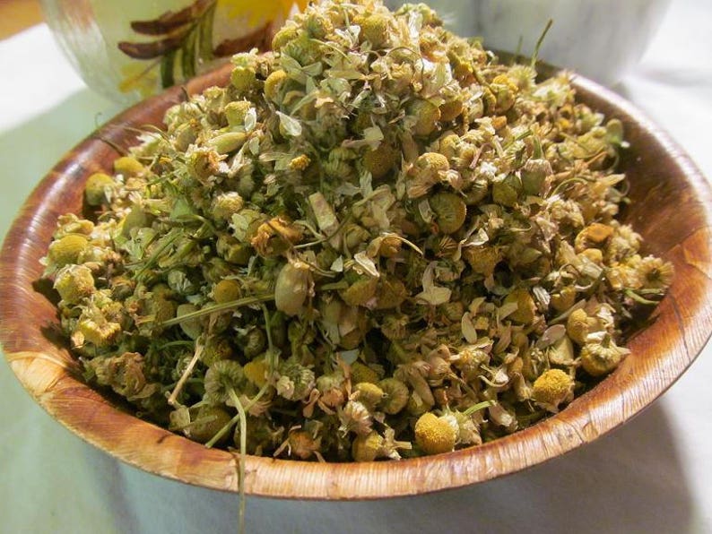 May include: A wooden bowl filled with dried chamomile flowers. The flowers are a mix of white petals and yellow centers.